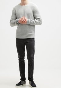 Muž stojící s dlaněmi spojenými, oblečený v světlém šedém tričku s dlouhým rukávem, černých roztrhnutých skinny džínách a černých teniskách s bílou podrážkou.