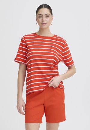 Femeie purtând un tricou cu mâneci scurte în dungi portocalii și albe și pantaloni scurți uni portocalii, cu cercei rotunzi și o expresie neutră.