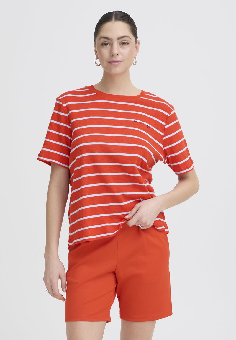 Frau trägt ein orange-weiß gestreiftes Kurzarmshirt und ein einfarbig oranges Shorts, mit Creolen und einem neutralen Ausdruck.