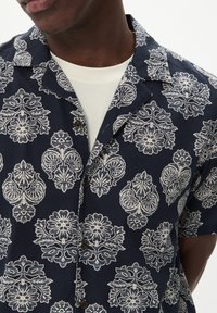 Camicia a maniche corte blu navy con un motivo floreale e paisley bianco, dotata di colletto a camicia e bottoni marroni, realizzata in tessuto leggero.