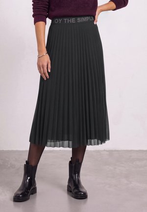 Le modèle porte une jupe midi plissée noire, un pull bordeaux, des collants transparents et des bottines noires. La ceinture de la jupe porte l’inscription « ENJOY THE SIMPLE ».