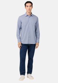 Camicia azzurro chiaro a maniche lunghe con colletto classico, indossata con jeans blu scuro e sneaker bianche, in piedi contro uno sfondo semplice.