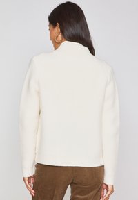 Maglione bianco a maglia con colletto alto e bordo a coste; presenta una finitura testurizzata. Abbinato a pantaloni in velluto a coste marroni.