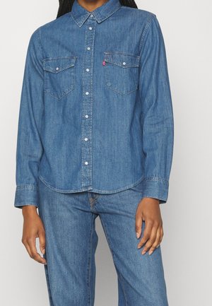 Femme portant une chemise en denim bleu à boutons avec deux poches poitrine et un jean en denim bleu assorti, sur un fond uni.