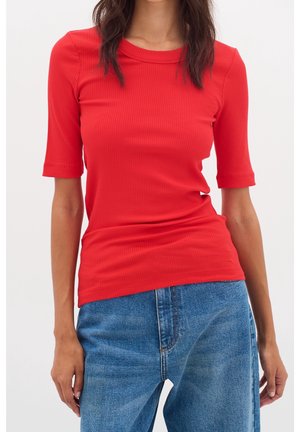 Vrouw draagt een aansluitend rood geribd kort mouw topje en blauwe denim jeans, staand tegen een effen witte achtergrond.