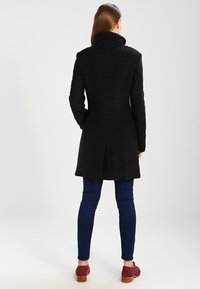 Donna in piedi di spalle, indossa un cappotto nero testurizzato, pantaloni fitted blu scuro e scarpe di camoscio rosse con tacchi in legno.