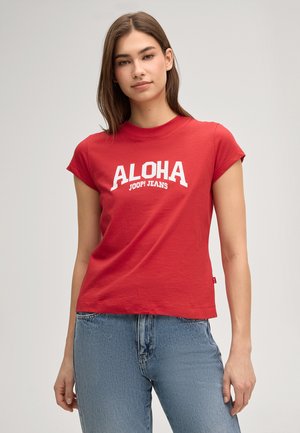Junge Frau mit langen dunklen Haaren, die ein rotes T-Shirt mit dem Schriftzug "ALOHA JOOP! JEANS" und blaue Jeans trägt, vor einem schlichten Hintergrund stehend.