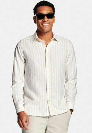 Lachende man met zwarte zonnebril, beige-wit gestreept overhemd met lange mouwen en lichte broek, met één hand in de zak.
