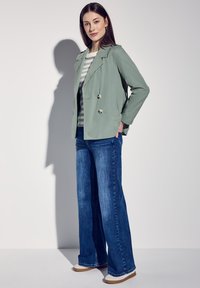 Zielony lekki płaszcz trench z podwójnymi guzikami, noszony na paskowanej koszuli, w połączeniu z szerokimi niebieskimi dżinsami i płaskimi beżowymi butami.