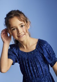 Blaues Smockkleid mit kurzen Puffärmeln, floralen Muster und gerafftem Ausschnitt. Das Model hat gewelltes Haar und fasst sich ans Ohr.