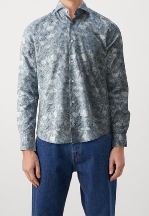Mand iført en langærmet skjorte med gråt og hvidt blomsterprint, kombineret med blå denimjeans.