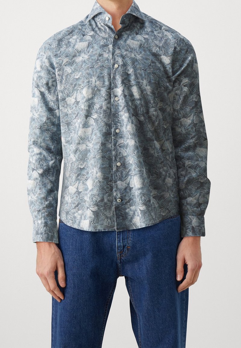 Homme portant une chemise à manches longues avec un motif floral gris et blanc, associée à un jean en denim bleu.