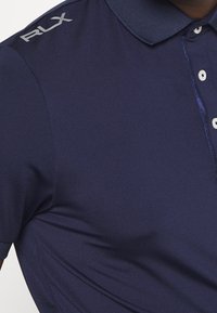 Mörkblå polo-shirt med en strukturerad krage, tre knappar i placket och en silverfärgad "RLX"-logotyp på axeln. Mjuk, slät tyg.