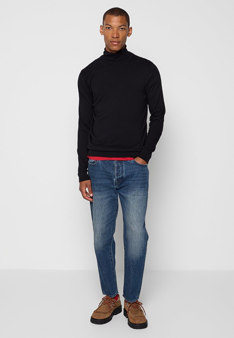 Mężczyzna w czarnym swetrze z golfem, niebieskich dżinsach i brązowych sznurowanych butach, stojący z jedną ręką w kieszeni.