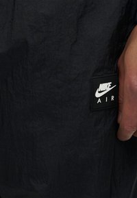 Μαύρο ύφασμα με ένα έμβλημα λογότυπου Nike Air και μέρος του χεριού ενός ατόμου που βρίσκεται δίπλα σε αυτό.