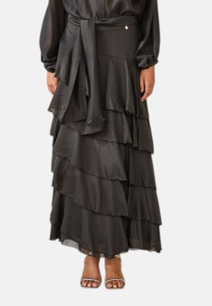 Gonna lunga nera a balze con volant e cintura in tessuto, indossata con sandali aperti con cinturini su una persona in piedi davanti a uno sfondo uniforme.