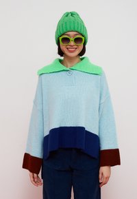 Personne souriante portant un bonnet en tricot vert, des lunettes de soleil vertes assorties et un pull color-block avec un col vert vif, des manches bleu clair, marine et marron.