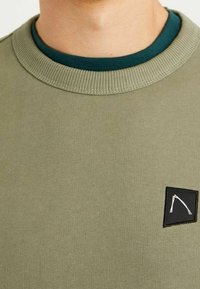 Sudadera verde oliva con cuello redondo acanalado, que presenta un pequeño parche negro con un detalle gráfico blanco en el pecho. Textura suave.