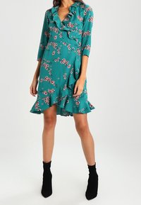 Türkises Blumenkleid mit Rüschensaum am Ausschnitt und Saum, Dreiviertelärmel und taillierter Passform. Getragen mit schwarzen Knöchelstiefeln.