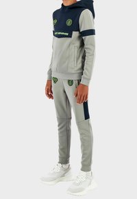 Grijs- en marineblauw trainingspak met capuchon, voorzien van groene logo's en accenten. Gemaakt van glad, rekbaar stof. Inclusief zijzakken en een aansluitend ontwerp.