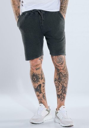 Mann, der schwarze strukturierte Shorts und weiße Sneakers trägt, Beine bedeckt mit verschiedenen detaillierten schwarzen Tattoos, einschließlich Rosen, Sonne und Schmetterling.