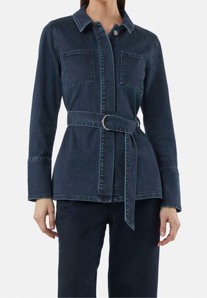 Kvinde iført mørkeblå denim-skjortejakke med frontlommer på brystet og matchende bælte bundet om taljen, kombineret med mørkeblå bukser.