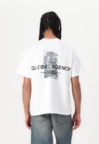 Bílý bavlněný tričko s grafickým potiskem palem a textem "GLOBAL AGENCY" na zadní straně, v uvolněném střihu a s krátkými rukávy.