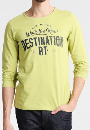 Hombre con camiseta de manga larga verde claro con texto negro "Walk the Road DESTINATION RT" y vaqueros oscuros, con la mano en el bolsillo.