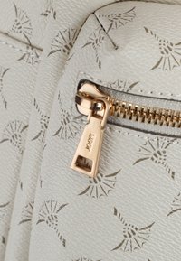 Wit textuurmateriaal met een subtiel patroon, voorzien van een ritssluiting en een trekband in gouden tint op een ritsvakdetail.