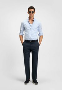 Mann mit Sonnenbrille, hellblau gestreiftem Hemd mit hochgekrempelten Ärmeln, dunklen Hosen, schwarzem Gürtel und schwarzen Schuhen mit weißen Sohlen, der steht.