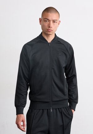 Juoda zip-up treniruočių striukė su guma apdirbtomis rankovėmis, minkšta tekstūra ir logotipo detale ant krūtinės. Yra stovintis apykaklė ir šoniniai kišenės.