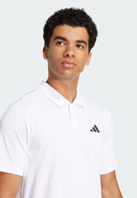 White polo shirt made of smooth fabric, featuring a classic collar, two buttons, and a black Adidas logo on the left chest.