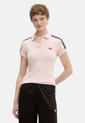 Persona joven con el pelo corto que lleva una camiseta polo rosa claro de Adidas con rayas negras y pantalones deportivos negros de Adidas.