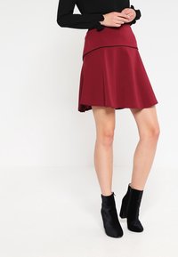 Jupe évasée rouge avec une taille ajustée et des détails en noir. Le mannequin porte des bottines noires. Tissu lisse avec une texture douce.
