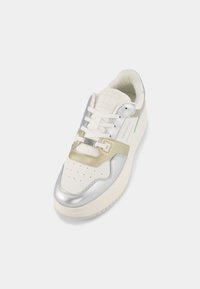 Basket blanche avec des empiècements argentés et beiges, lacets blancs et perforations sur l'avant, montrée en vue plongeante oblique sur fond blanc.