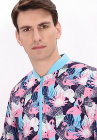 Bloemen bomberjack met een marineblauwe achtergrond, levendige roze flamingo's en een lichtblauwe rits met een ribgebreide kraag.
