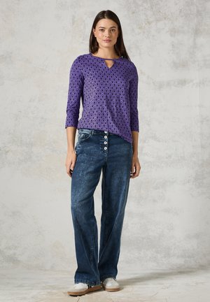 Lila gemusterte Bluse mit V-Ausschnitt und Dreiviertelärmeln, kombiniert mit blauen Jeans im weit geschnittenen Stil mit Knopfakzenten und weißen Sneakers.