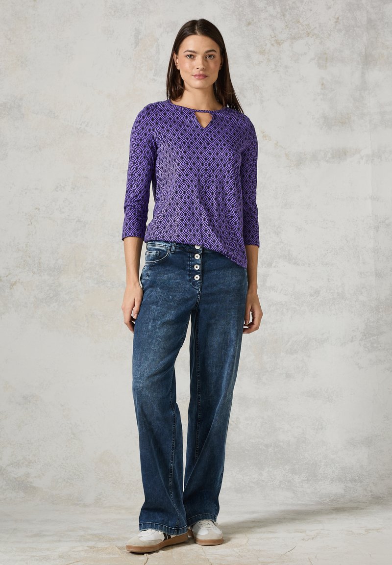 V-pääntie ja kolme neljännestä hihasta varustettu violetti kuvioitu yläosa, yhdistettynä sinisiin leveälahkeisiin farkkuihin, joissa on nappikoristeita, ja valkoisiin tennareihin.