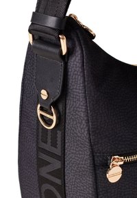 Sac à épaule en tissu noir avec une surface texturée, accents en métal doré, sangle réglable et détails de poches zippées.