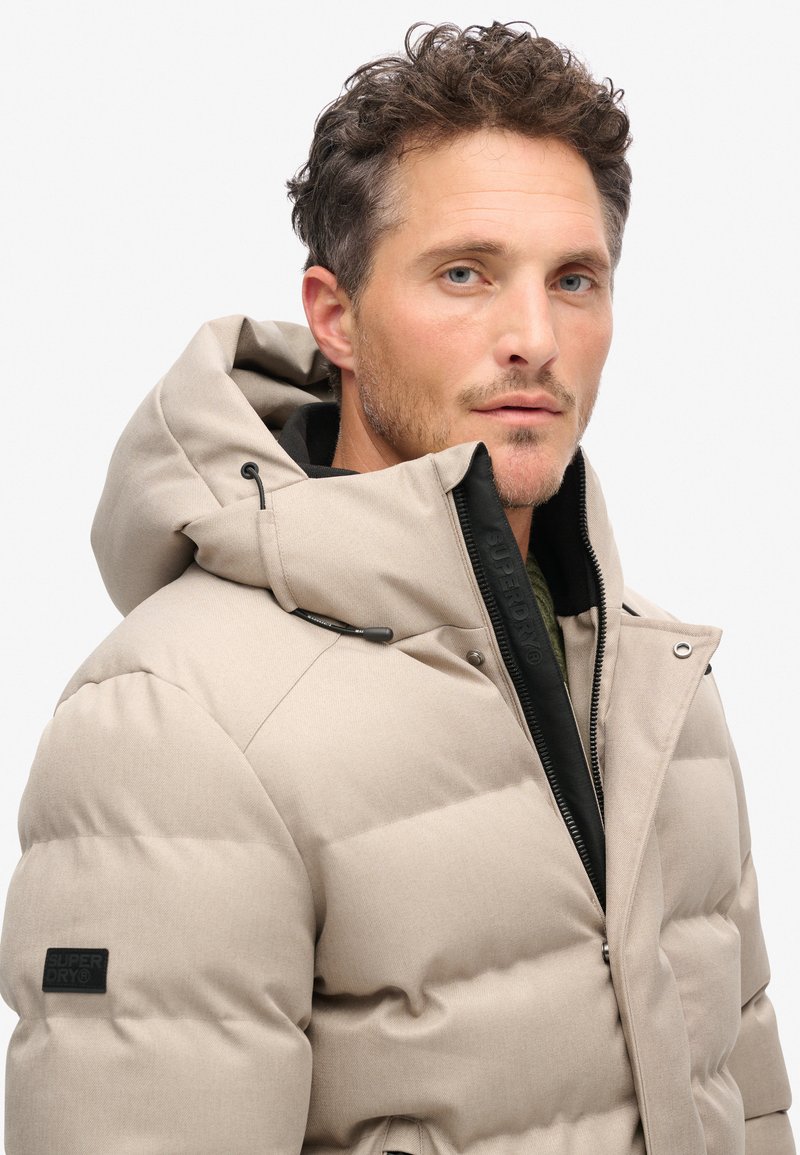 Beige gewatteerde jas met een capuchon, voorzien van doorgestikte panelen, een zwarte rits en een merklogo op de mouw. Gladde textuur, middellange ontwerp.