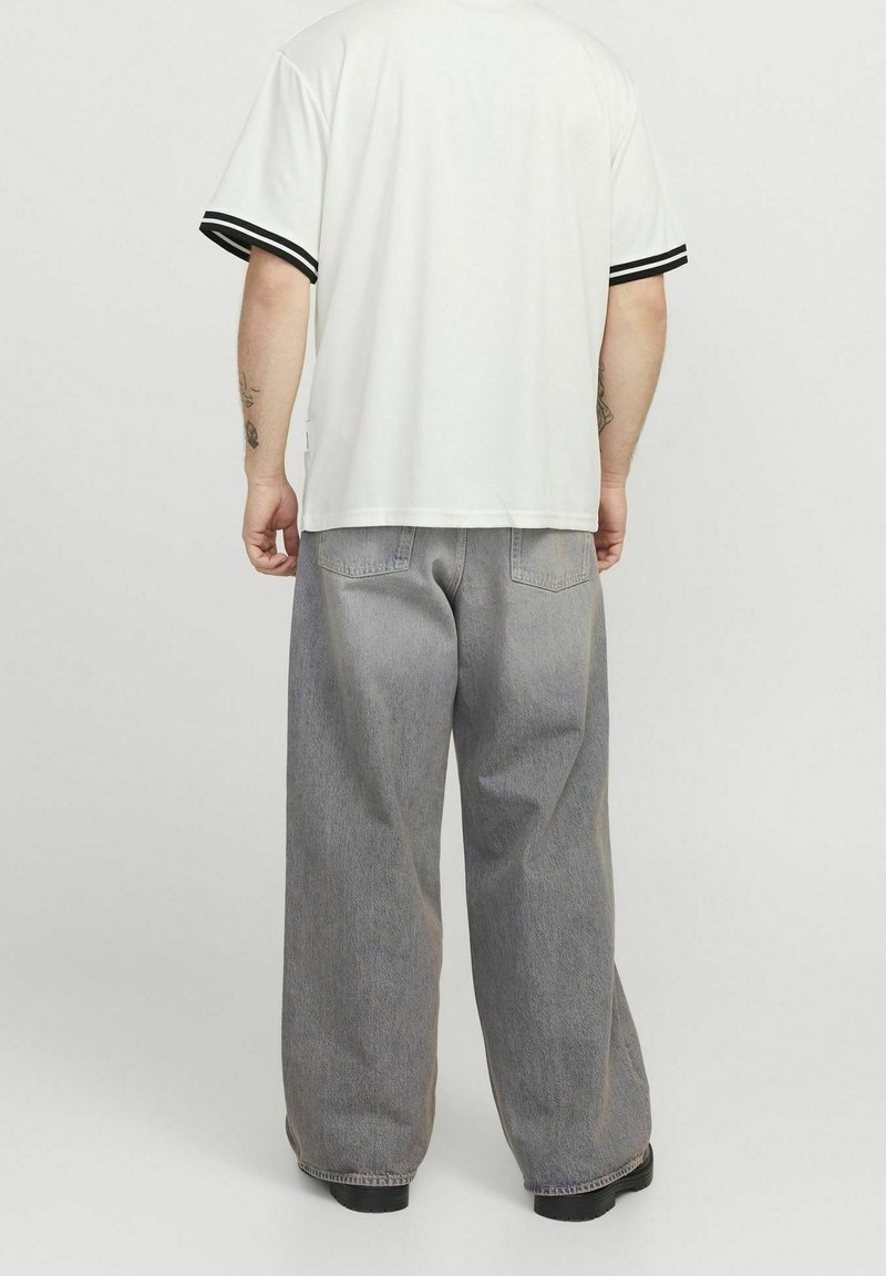 Bílá košile s krátkým rukávem a černými pruhovanými manžetami, v kombinaci s volnými světle šedými džíny. Tkanina vypadá hladce a ležérně.