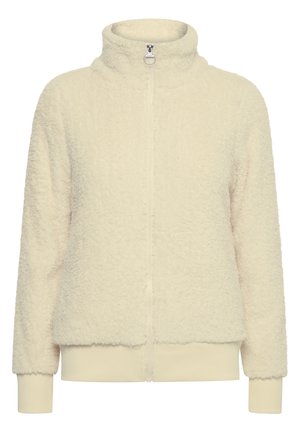 Person iført cremefarvet fleecejakke med lynlås, mørkeblå stramme jeans og beige snørestøvler står foran en ensfarvet baggrund.