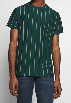 Mężczyzna w ciemnozielonej koszulce z krótkim rękawem, z cienkimi pionowymi żółtymi paskami i niebieskich dżinsach, stojący z ramionami swobodnie opuszczonymi wzdłuż ciała.