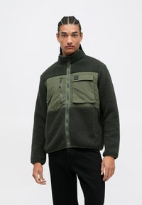Fleece jacket in dark green with a lightweight fabric upper and chest pocket. Features a high collar and zip closure. Textured, soft material.