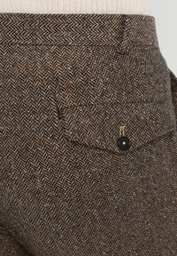 Brown herringbone-patterned trousers with a black button closure and patch pocket. Soft texture with visible weave detailing.
