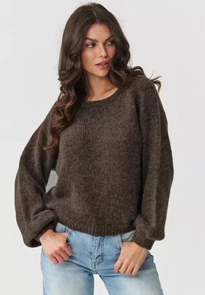 Brązowy oversize'owy sweter z dzianiny o ryflowanej fakturze, okrągłym dekolcie i obszerności rękawów, w połączeniu z jasnoniebieskimi dżinsami.