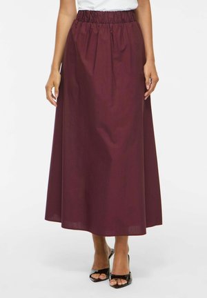 Femme portant un haut blanc, une jupe bordeaux longue jusqu'aux chevilles avec une ceinture élastique et des sandales à talons noires ouvertes, debout devant un fond blanc.