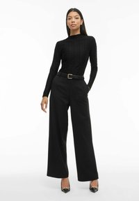 Mustat teksturoidut pitkähihaset paidat, leveät mustat housut vyöllä. Mustat kärjelliset korot, joissa on kultaketjuyksityiskohta. Yksinkertainen ja yksivärinen muotoilu.