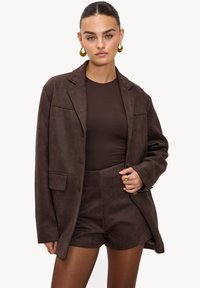 Blazer en daim marron avec une coupe ajustée, une poche poitrine à simple rabat et une fermeture à boutons, accompagné d'un haut marron assorti et d'un short.