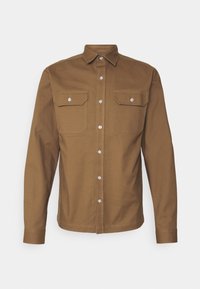 Chemise marron à manches longues avec boutons, deux poches poitrine avec rabats boutonnés et col pointu sur fond clair.